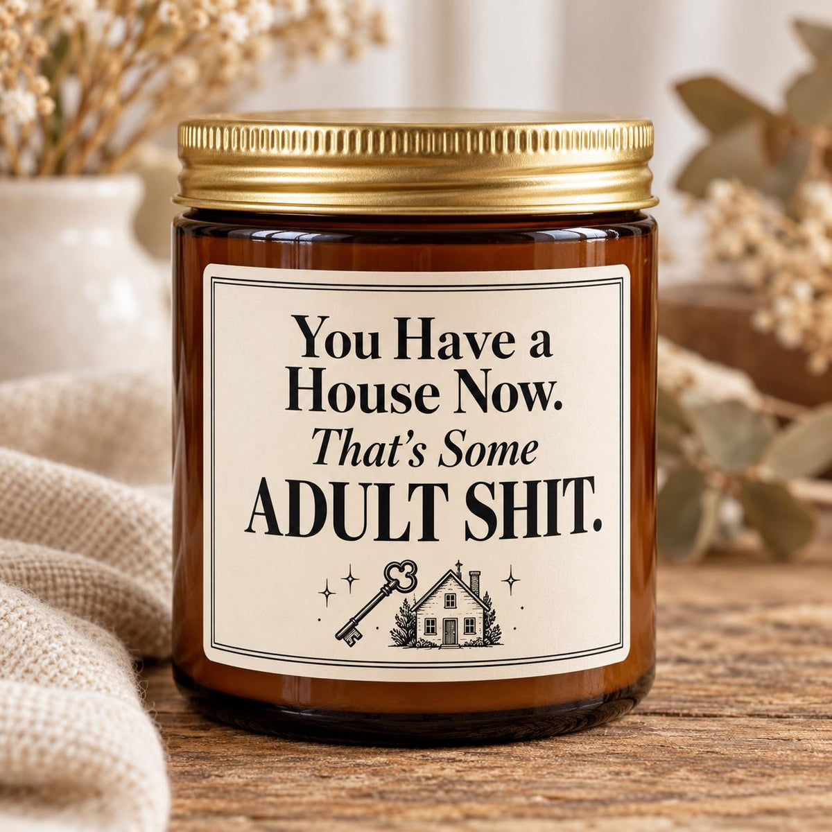An amber glass jar candle with a gold metal lid and a cream label that reads, 'You Have a House Now. That's Some ADULT SHIT.
