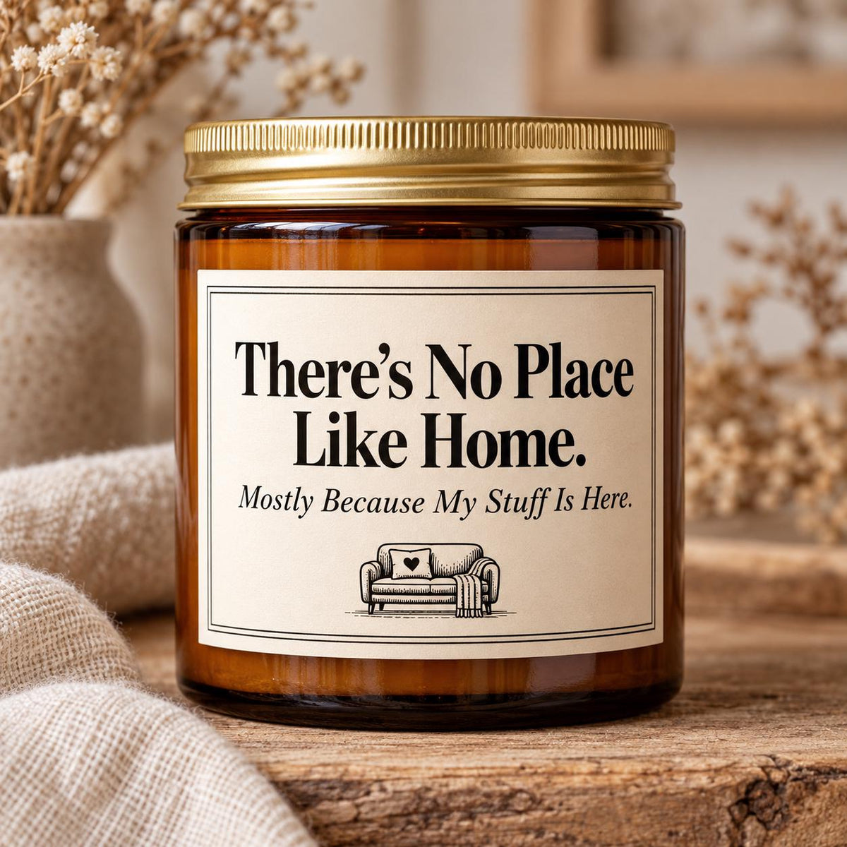 An amber glass jar candle with a gold metal lid and a cream label that reads, 'There's No Place Like Home. Mostly Because My Stuff Is Here.