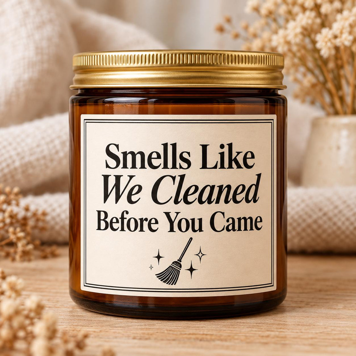 An amber glass jar candle with a gold metal lid and a cream label that reads 'Smells Like We Cleaned Before You Came' with a broom and sparkle illustration, set on a wooden surface with soft neutral background decor.