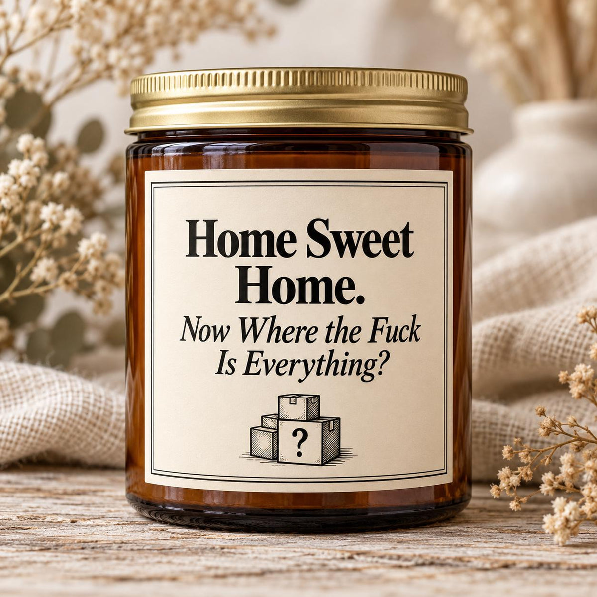 An amber glass jar candle with a gold metal lid and a cream-colored label that reads 'Home Sweet Home. Now Where the Fuck Is Everything?' with a small box illustration.