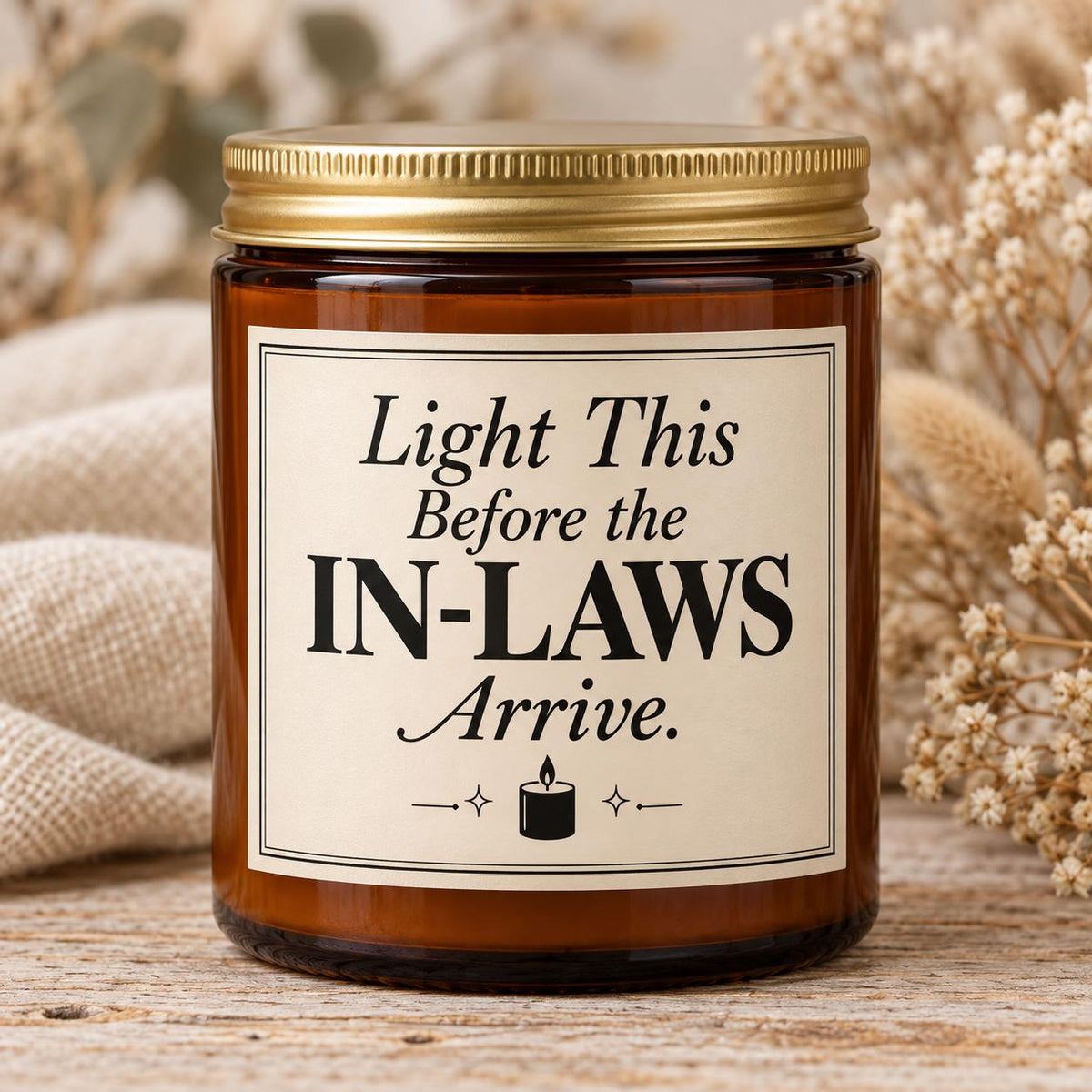 An amber glass jar candle with a gold metal lid and a cream label that reads 'Light This Before the IN-LAWS Arrive.' The candle is styled on a rustic wooden surface with burlap fabric and dried flowers in the background.