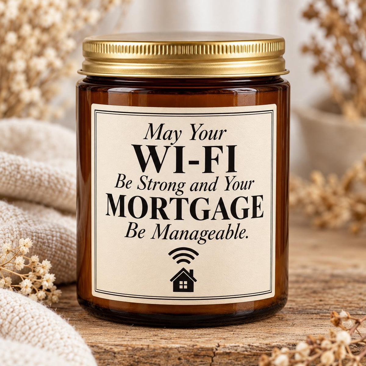 An amber glass jar candle with a gold metal lid and a cream label that reads, 'May Your WI-FI Be Strong and Your MORTGAGE Be Manageable.' The jar is styled on a rustic wooden surface with beige fabric and dried flowers in the background.