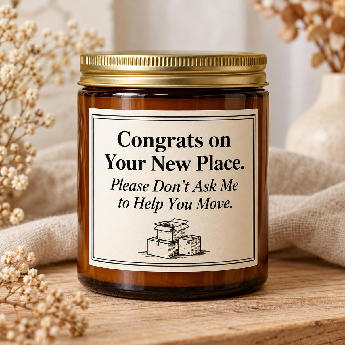 An amber glass jar candle with a gold metal lid and a cream label that reads 'Congrats on Your New Place. Please Don't Ask Me to Help You Move.