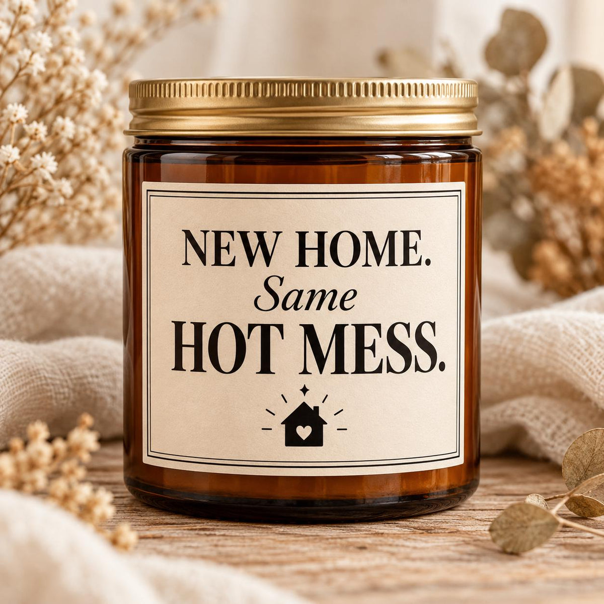 An amber glass jar candle with a gold metal lid and a cream label that reads 'NEW HOME. Same HOT MESS.