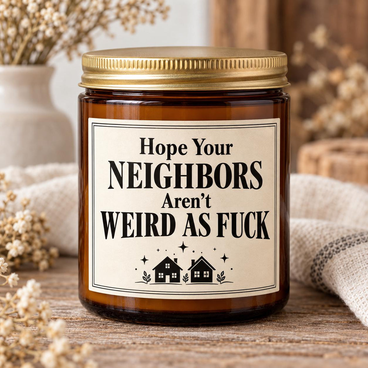 An amber glass jar candle with a gold metal lid and a cream label that reads 'Hope Your Neighbors Aren't Weird As Fuck' with small house illustrations, styled on a wooden surface with dried flowers and a burlap cloth in the background.
