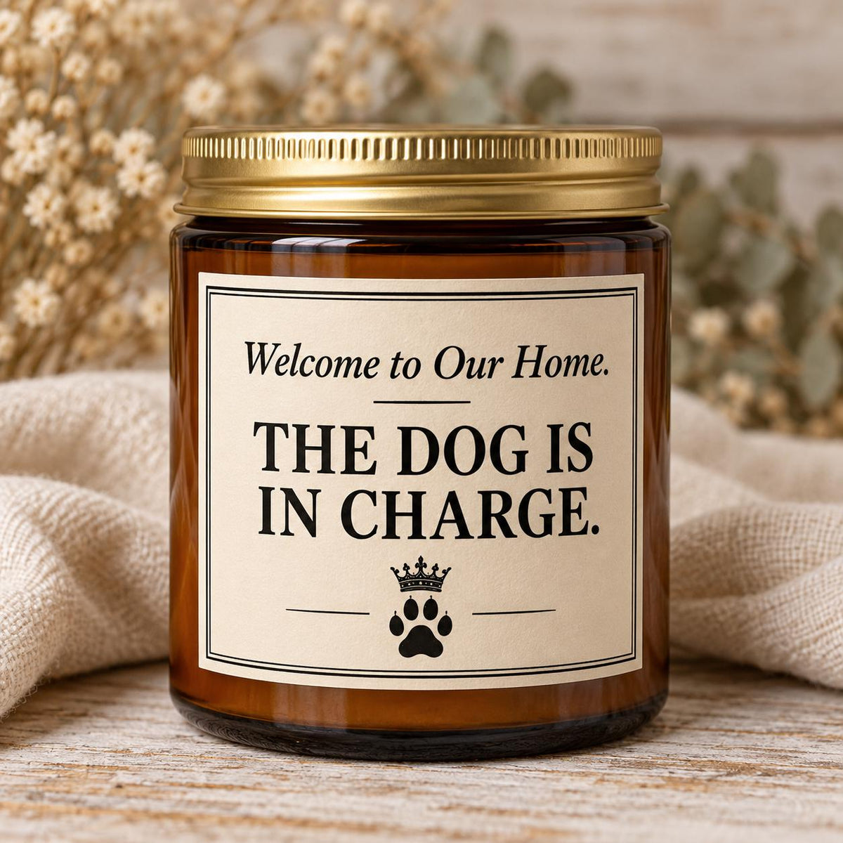 An amber glass jar candle with a gold metal lid and a cream label that reads 'Welcome to Our Home. THE DOG IS IN CHARGE.